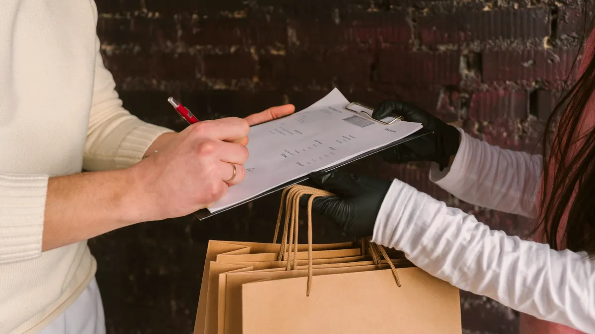 A courier in black gloves holds out a clipboard and a row of kraft takeout bags while a customer signs the delivery slip.