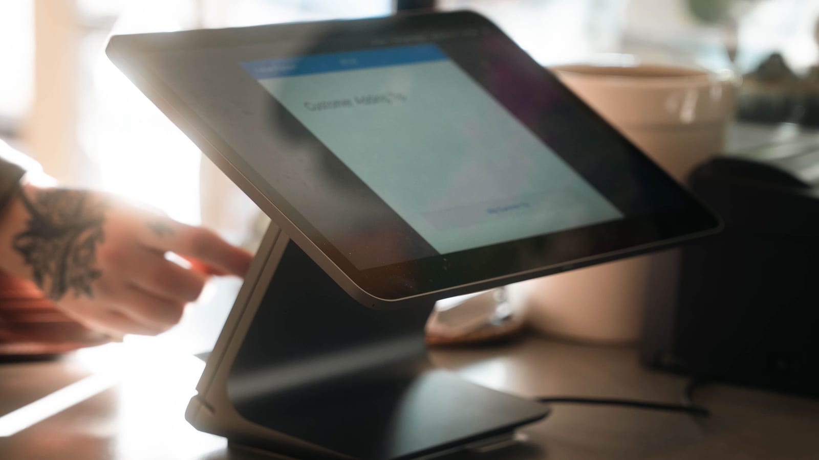 A hand reaches toward a tablet register on a café counter, its screen open to add a customer.