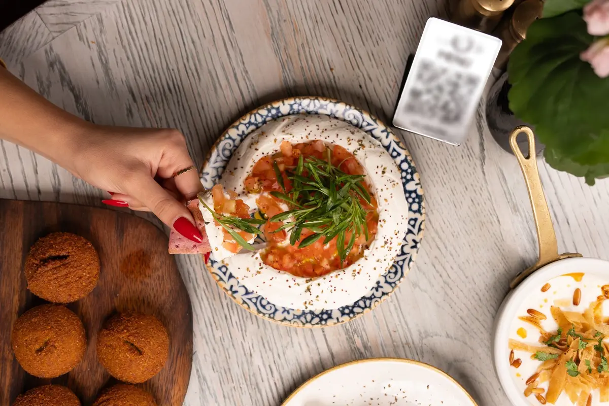 A hand dips bread into a plate of hummus beside falafel and a small table card with a QR code.