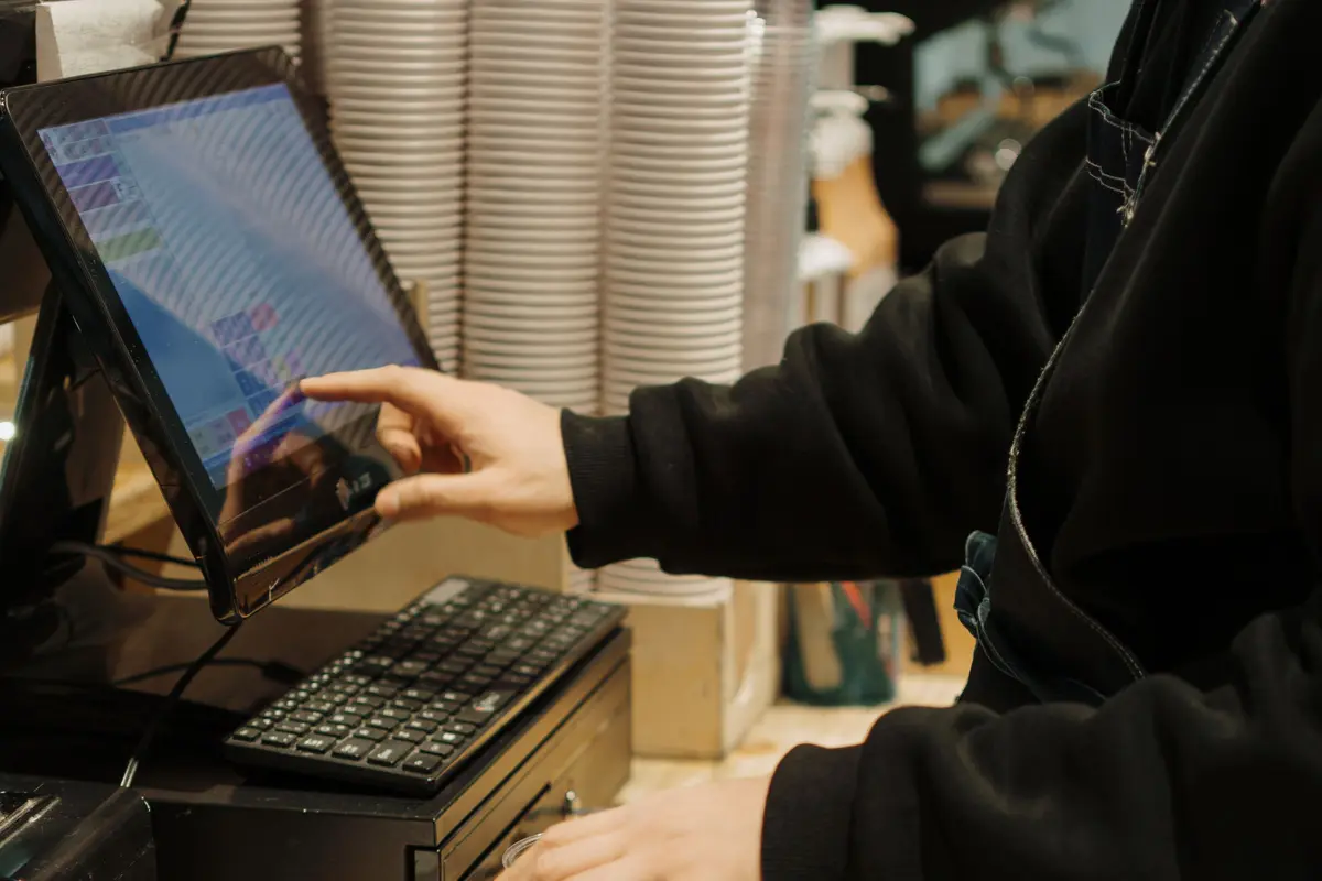A cashier’s hand taps an item into a touchscreen register at a café counter, stacked cups behind.