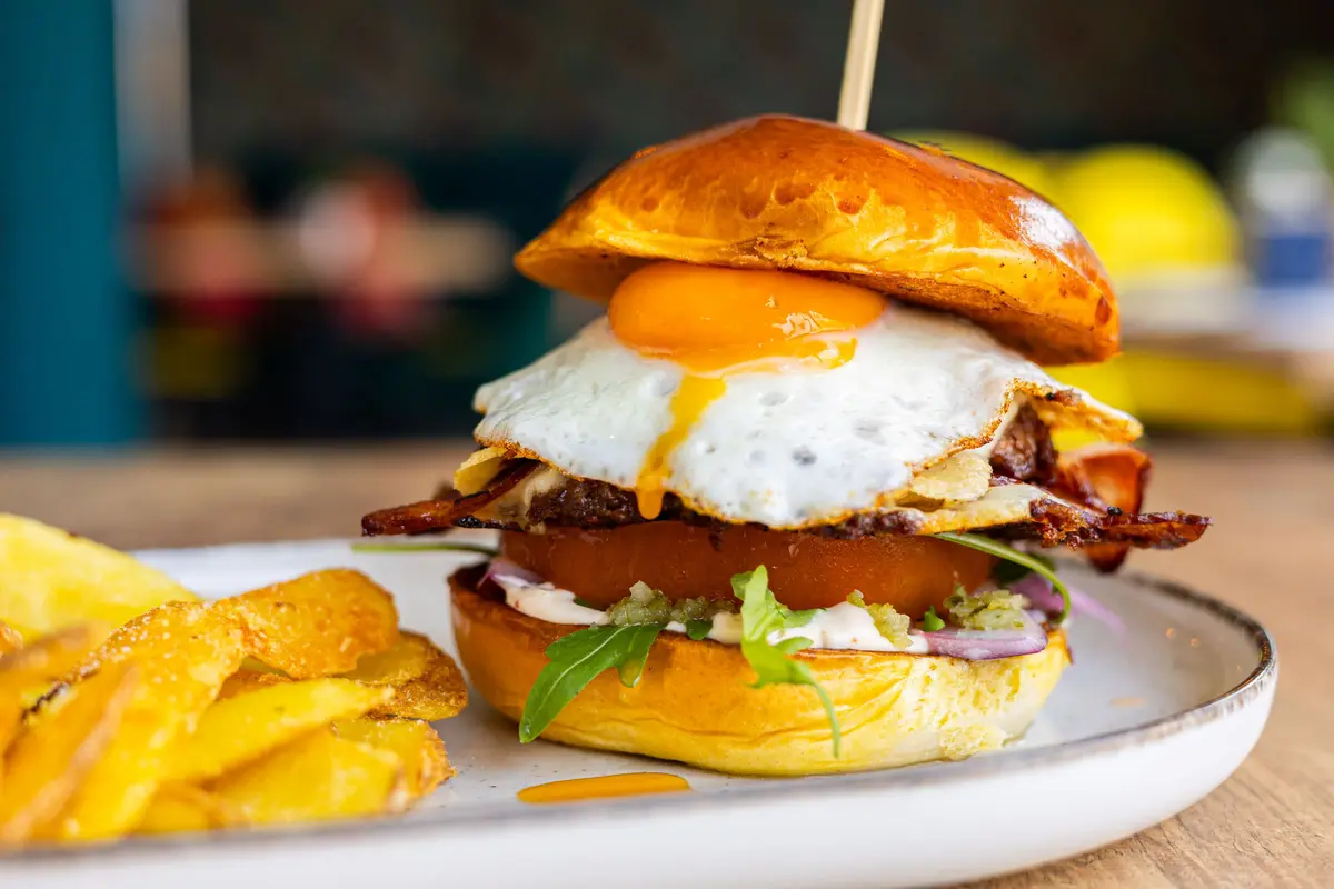 A burger with a fried egg, bacon and tomato sits on a white plate beside crisp potatoes, the yolk and bun in sharp focus.