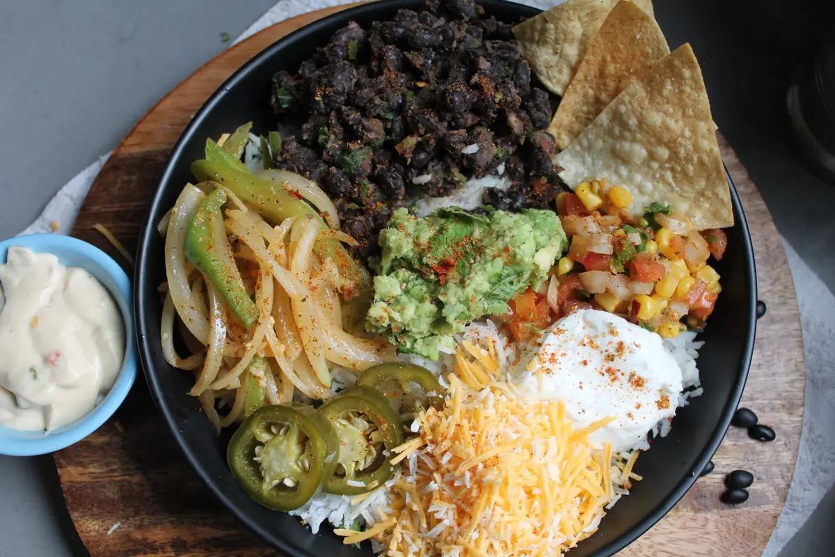 A rice bowl topped with black beans, guacamole, salsa, cheese and peppers, with tortilla chips on the side.
