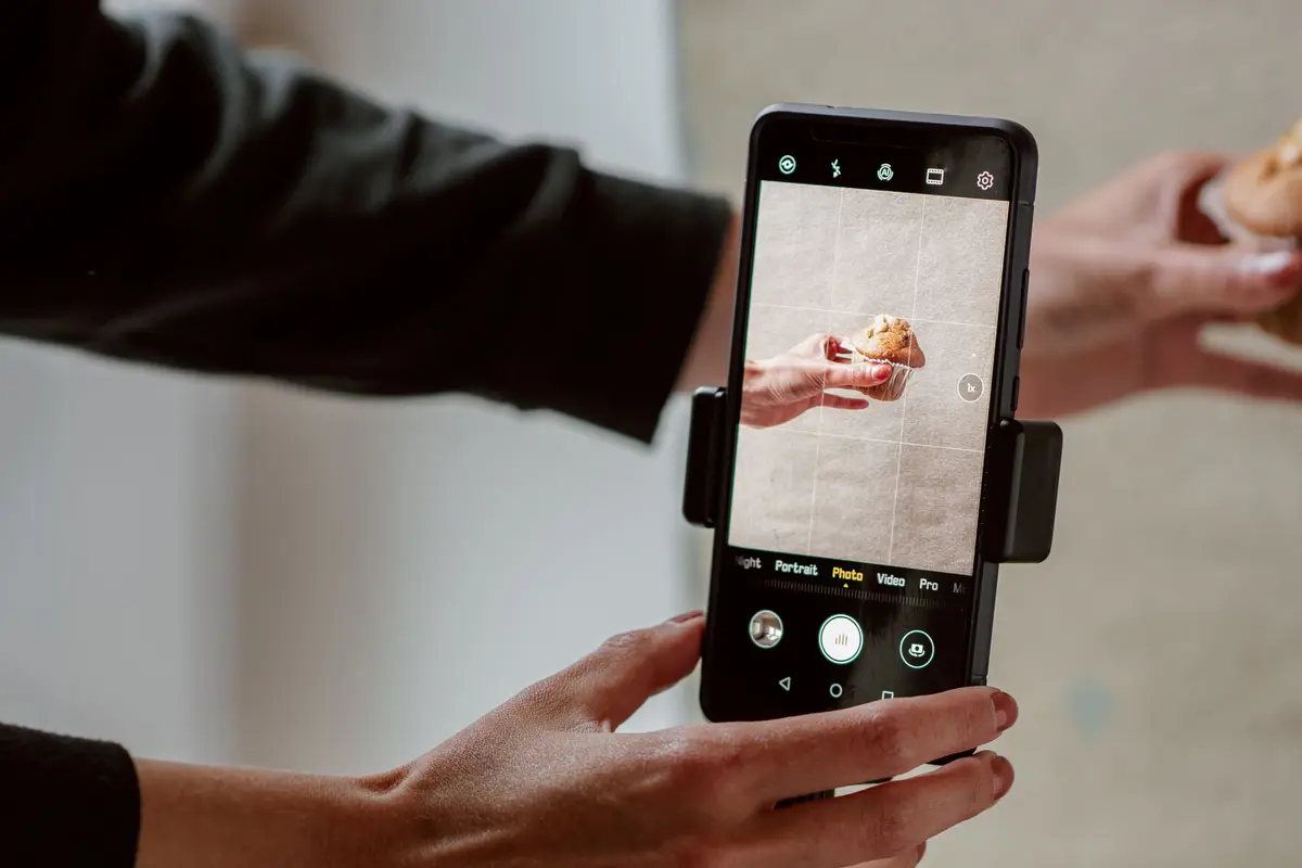A phone in a small mount photographs a muffin held up by hand at a table.