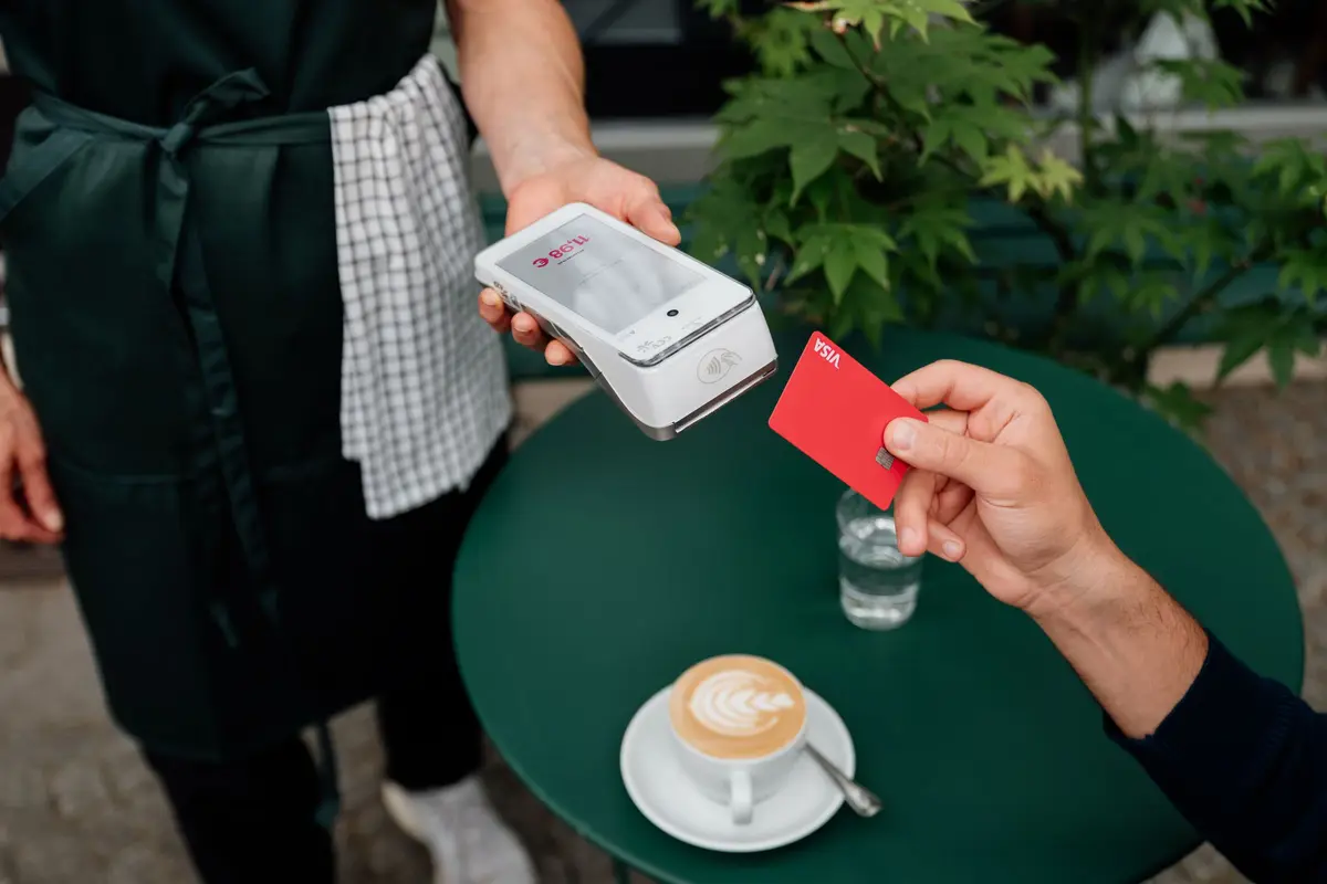 A server holds out a card reader while a guest taps a card over a cappuccino at an outdoor cafe table.