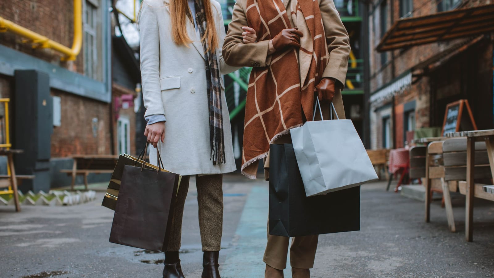 Two shoppers in winter coats carry paper shopping bags down a brick street past outdoor café tables.