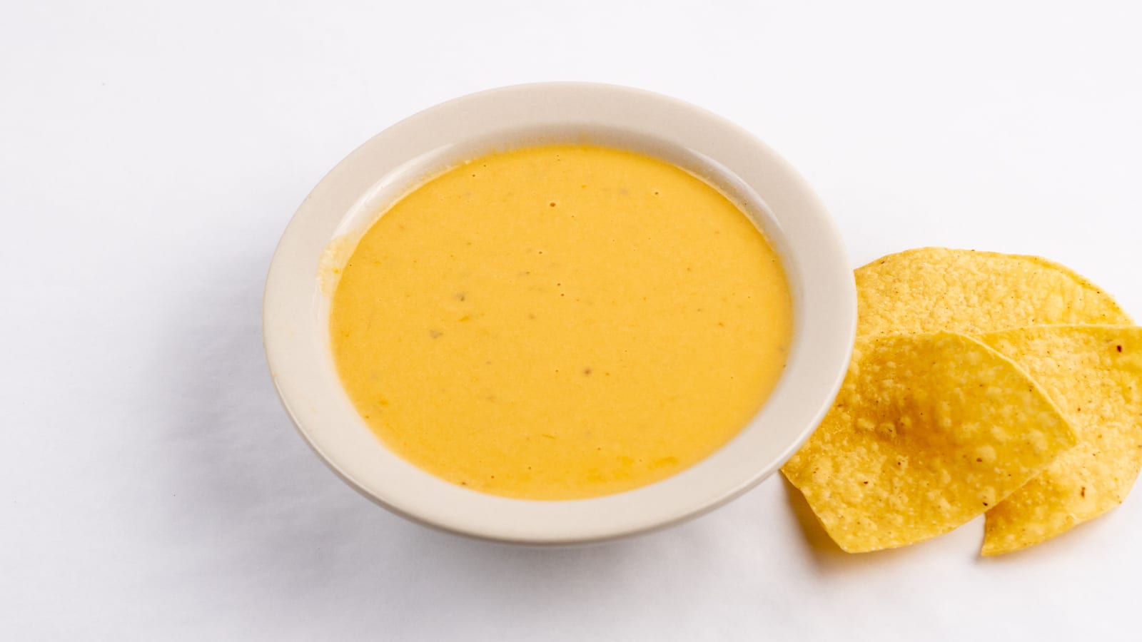 A white bowl of melted queso sits on a white table beside a few tortilla chips.