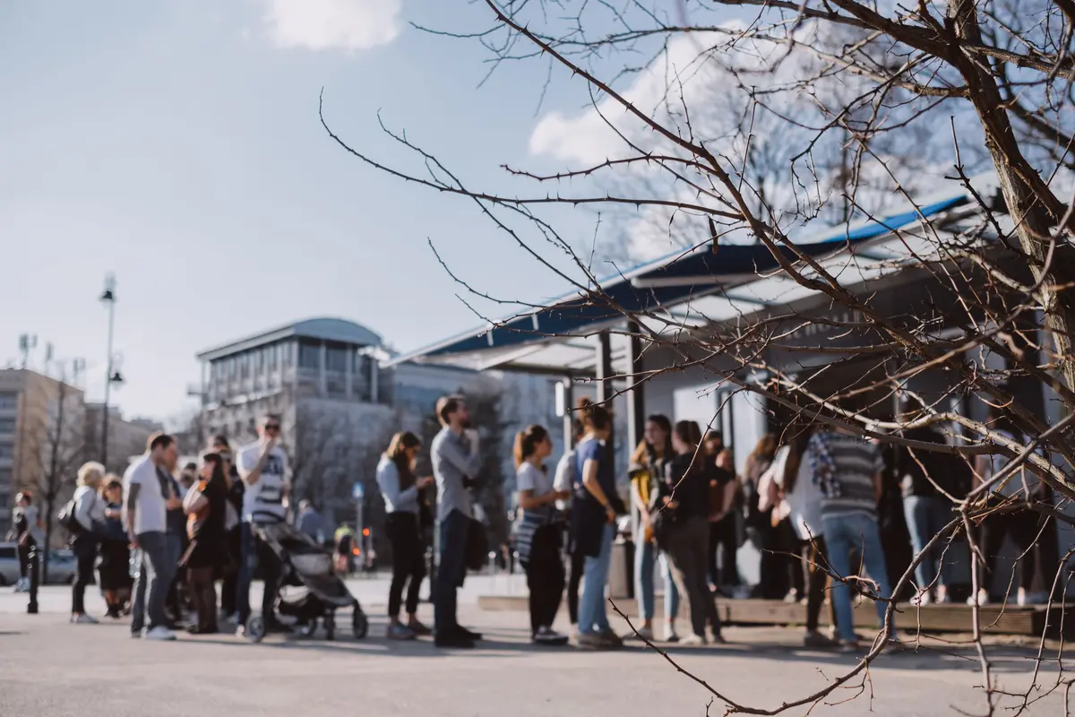 A long line of people waits outside a small pavilion on a bright day, out of focus behind bare branches.