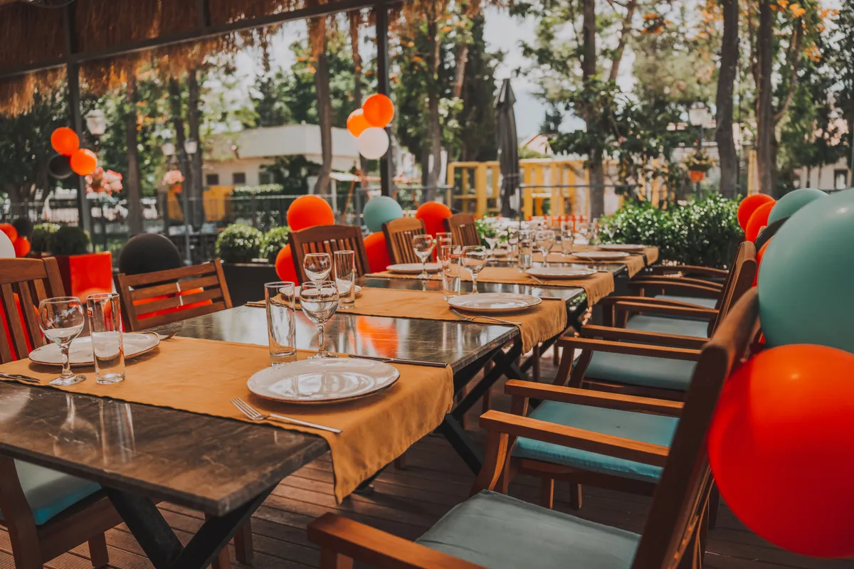 A long patio table is set with plates and glasses, with orange and teal balloons tied to the chairs.