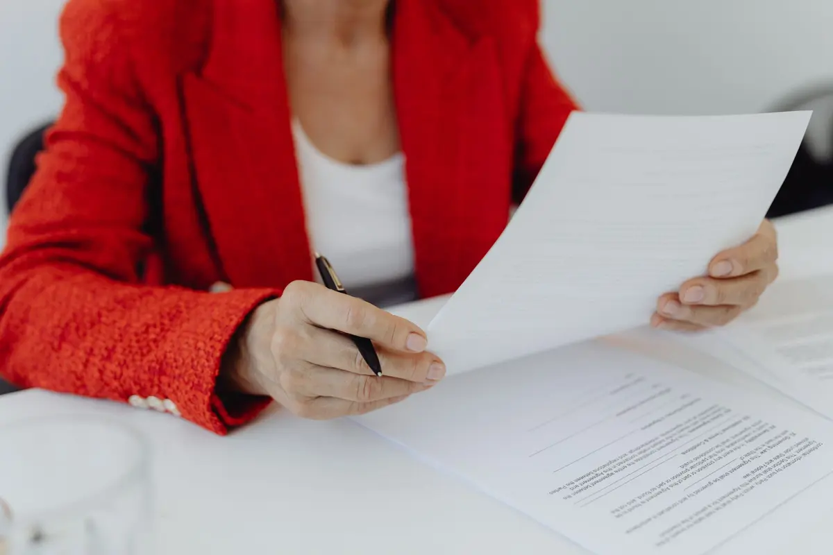 A person in a red jacket reads a printed page at a desk, a pen in hand and more pages beside it.
