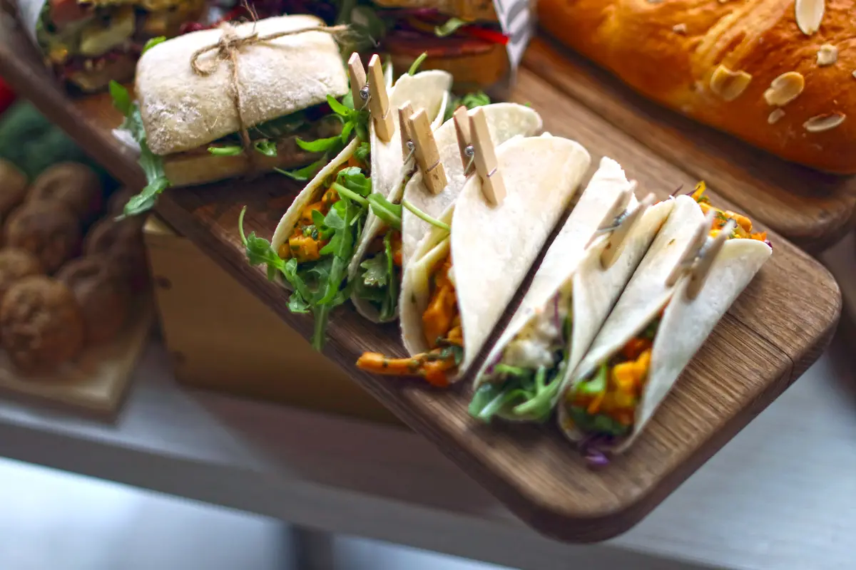 Soft tacos held with wooden pegs and a sandwich on a wooden catering board, the kind of tray a store sends to an office nearby.