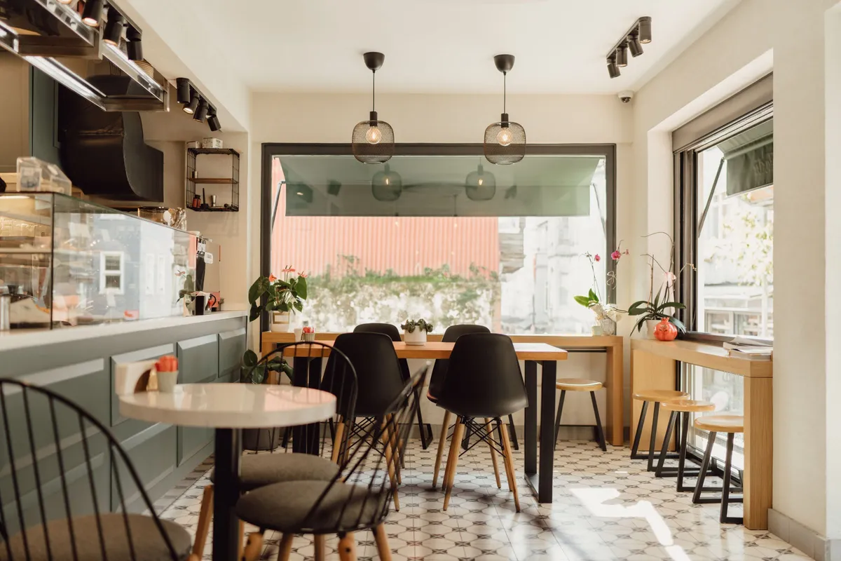 A small, sunlit cafe with empty black chairs, a tiled floor and a pastry case, before the first guests arrive.