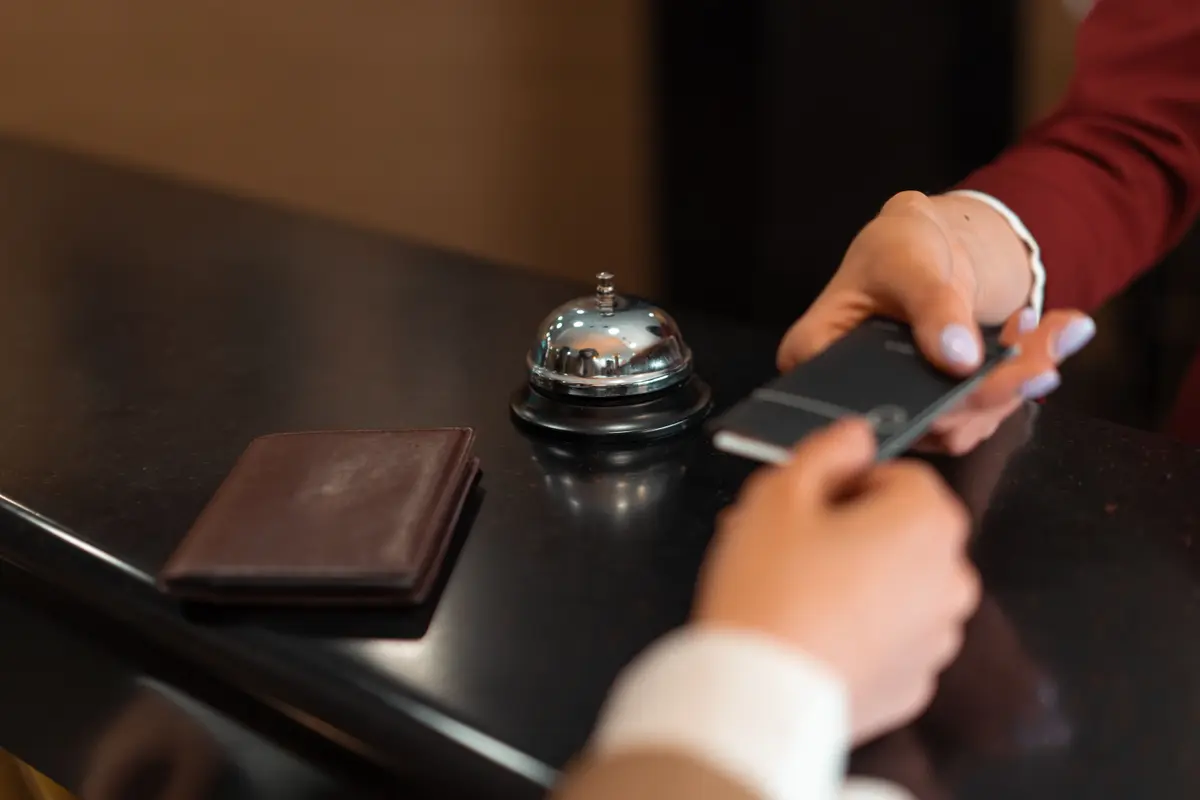 A staff hand in a red sleeve passes a small dark card across a front desk to a guest, a service bell and a wallet beside them.