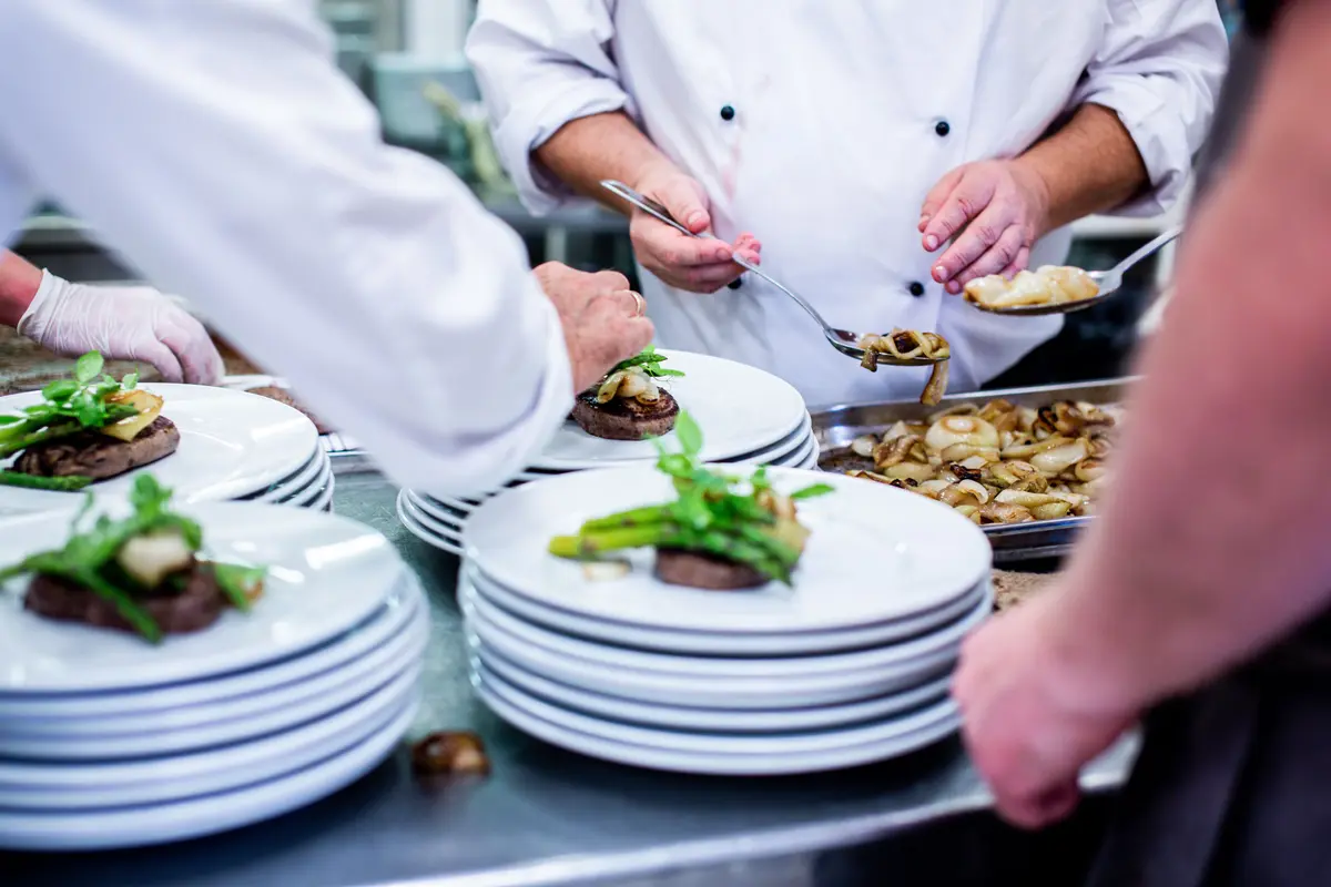 Chefs spoon onions onto plated steak and asparagus stacked at a restaurant kitchen pass while a server waits at the side.