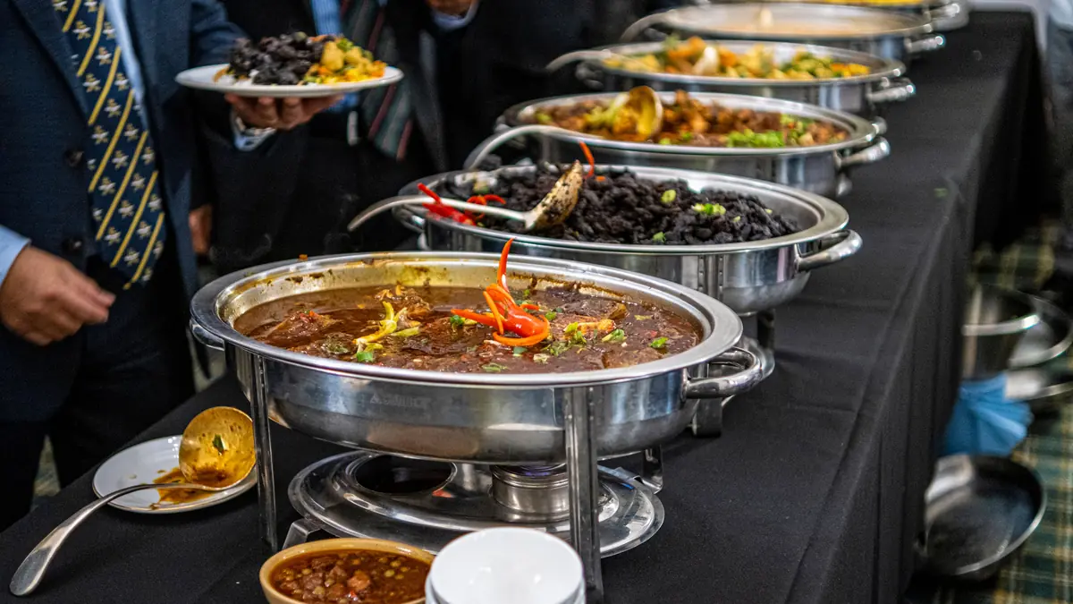 Guests in suits fill plates from a row of steel chafing dishes on a black-draped buffet table.