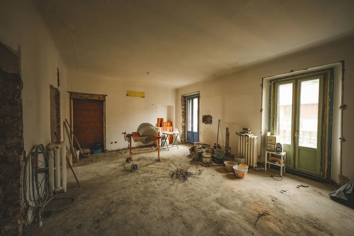 A bare room under renovation, with a cement mixer, stacked bricks and buckets on a dusty concrete floor and light coming through tall glass doors.