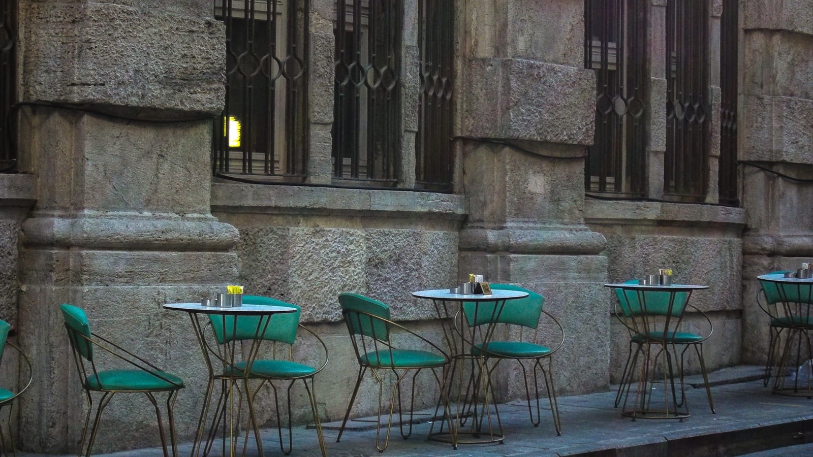 Empty green cafe chairs and small round tables line a stone sidewalk outside an old building.